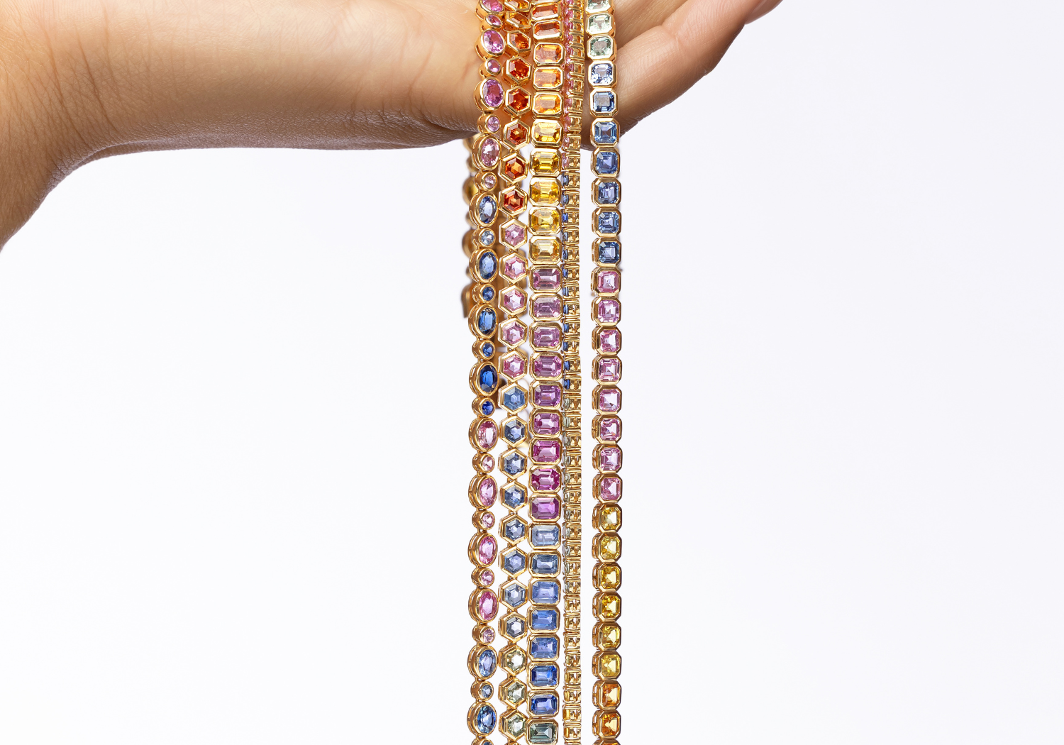 A hand holding a cascade of multi-colored sapphire tennis necklaces in yellow gold: blue, pink, yellow, orange, and green.