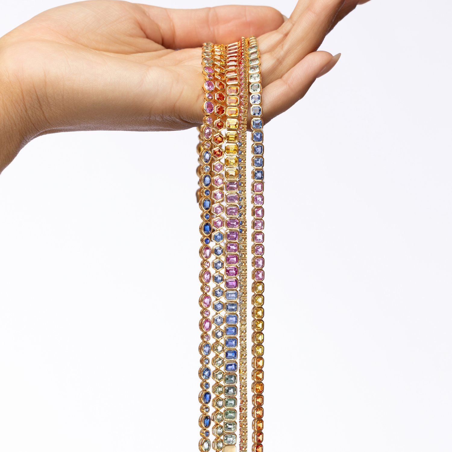 A hand holding a cascade of multi-colored sapphire tennis necklaces in yellow gold: blue, pink, yellow, orange, and green.