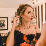Courtney McBroom wearing black onyx and mixed-shaped diamond teardrop earrings, paired with a black floral dress and matching black gemstone necklace in a softly lit dressing room.