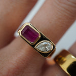 Striking east-west ring crafted in 14K yellow gold, featuring a bezel-set emerald-cut ruby and a sparkling pear-shaped diamond, exuding sophistication and timeless charm as a bold statement piece. Front facing, close up, on model.
