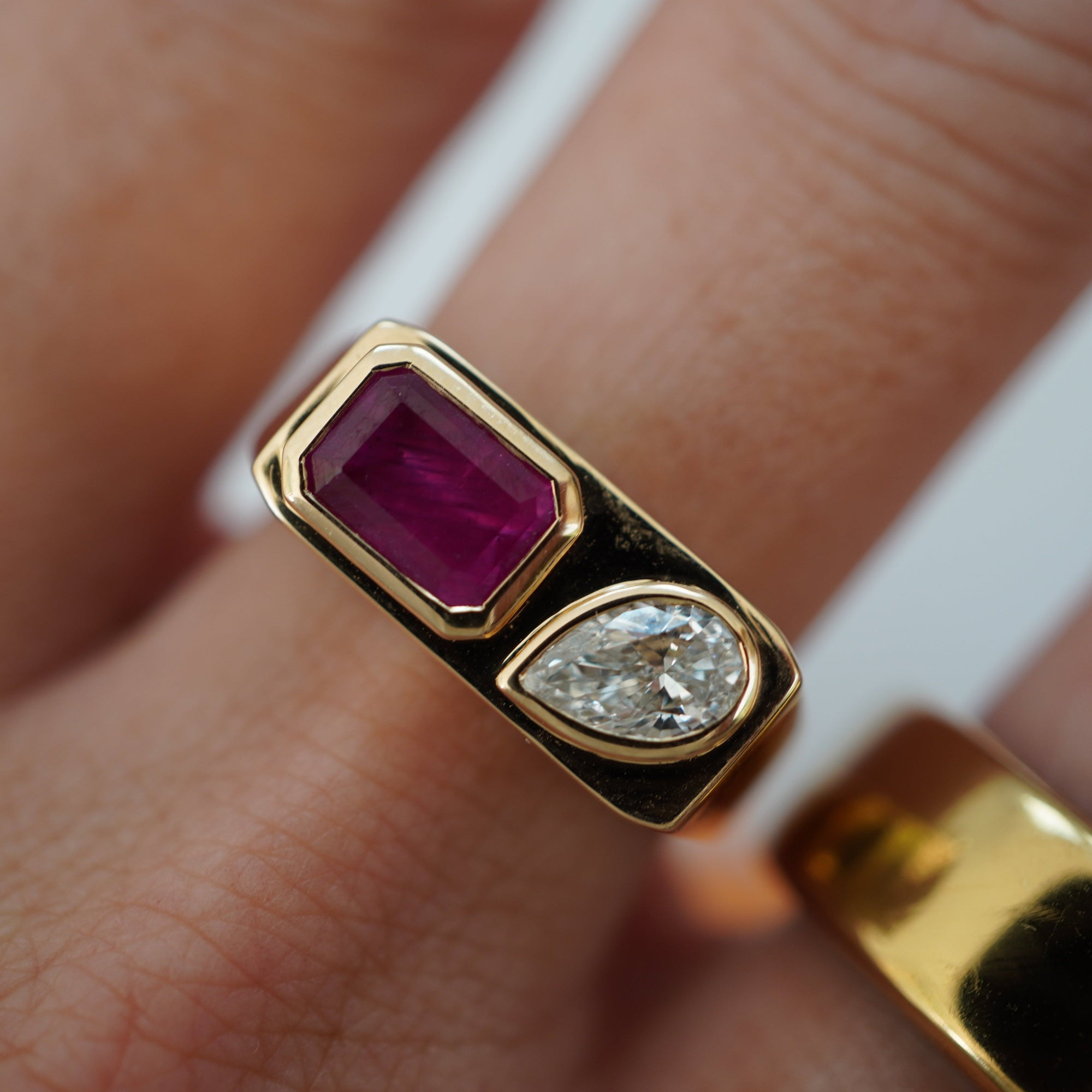Striking east-west ring crafted in 14K yellow gold, featuring a bezel-set emerald-cut ruby and a sparkling pear-shaped diamond, exuding sophistication and timeless charm as a bold statement piece. Front facing, close up, on model.