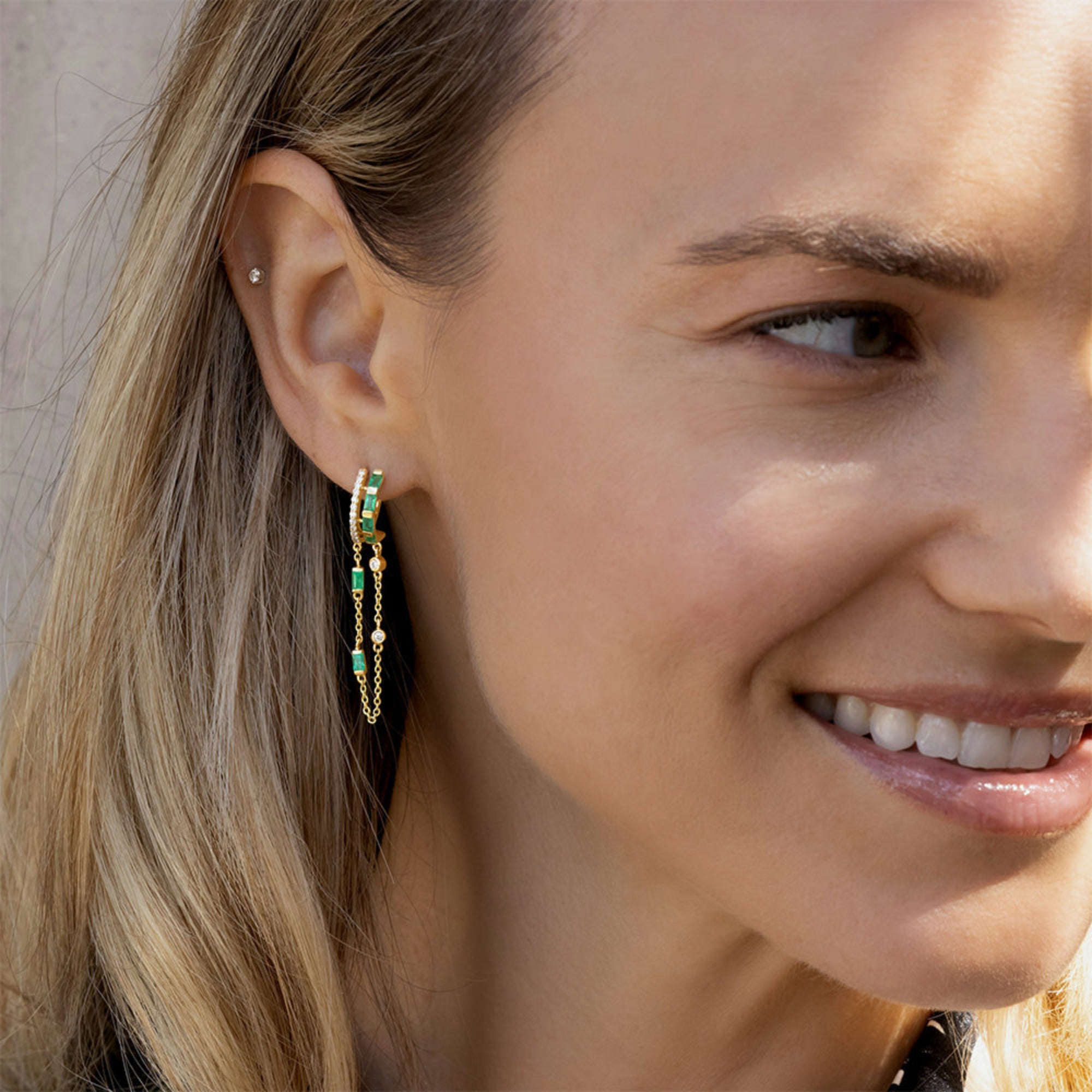 Captivating double hoop earrings featuring shimmering diamonds and vibrant emeralds in a double huggie design, accentuated by delicate dangling chains for a blend of classic sophistication and modern allure. On model in right ear. 