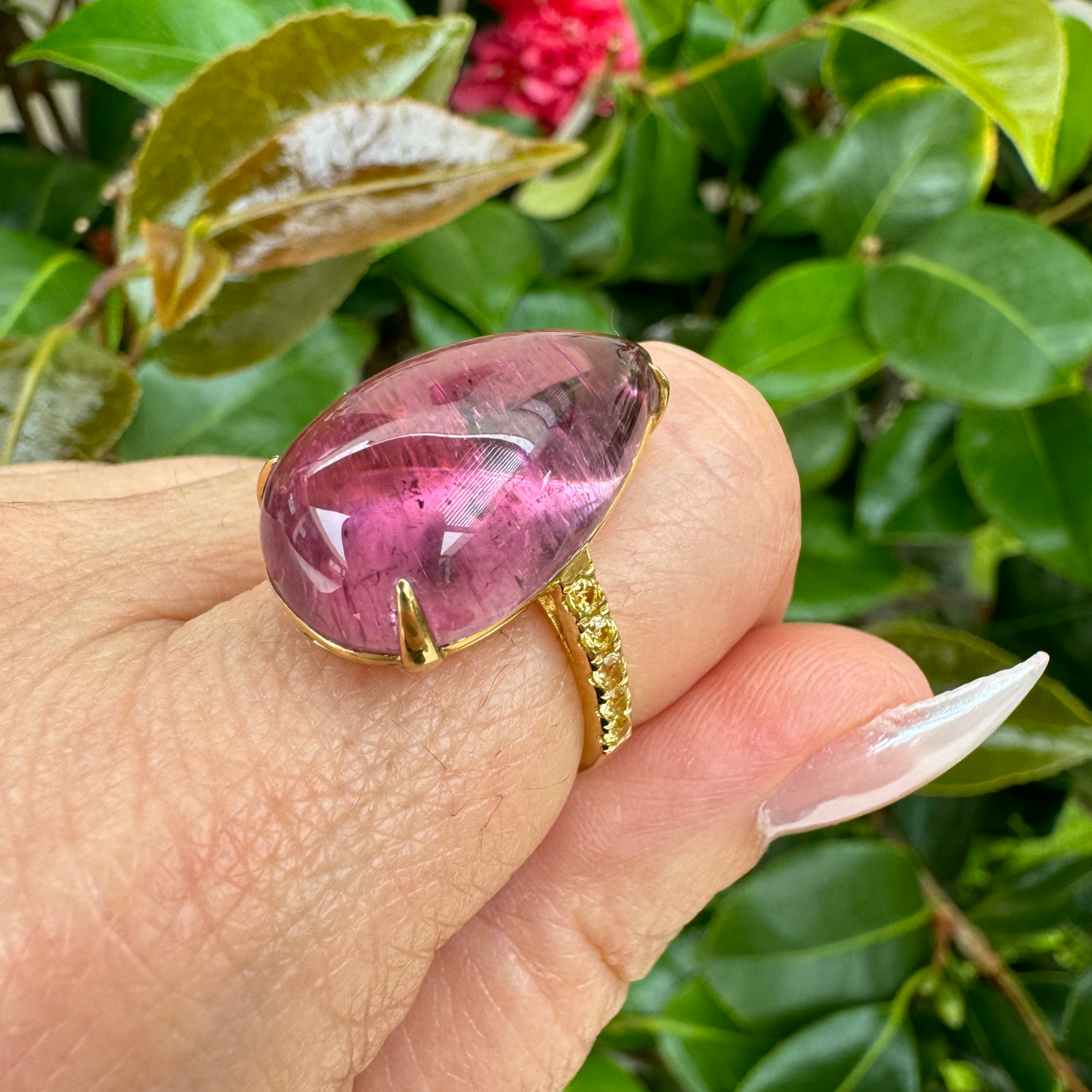 Exquisite 18K yellow gold ring featuring a breathtaking hand-carved pink tourmaline cabochon with a luminous glow, accented by vibrant yellow sapphires for a striking contrast of warmth and brilliance. On model hand, side angle with band and stone.