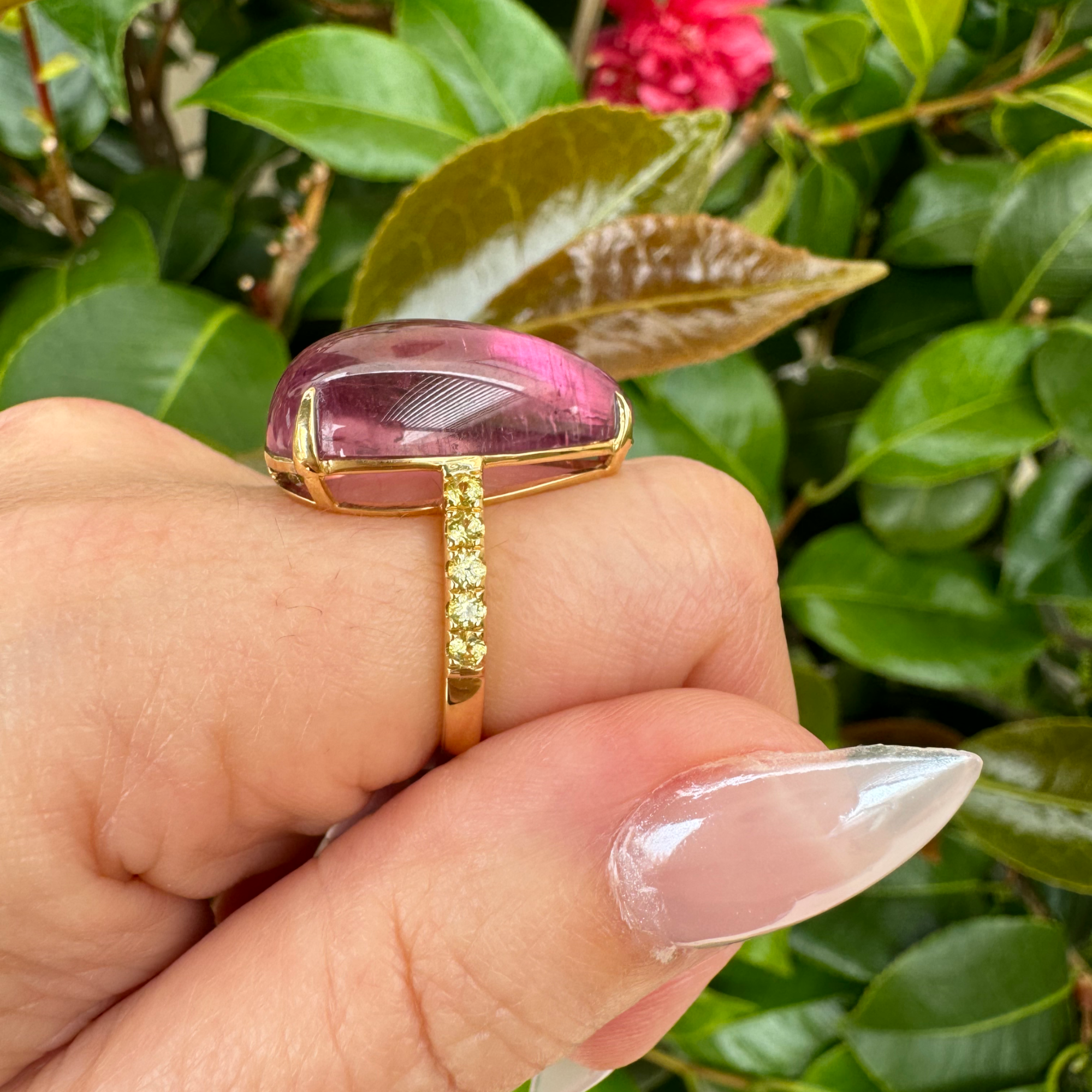 Exquisite 18K yellow gold ring featuring a breathtaking hand-carved pink tourmaline cabochon with a luminous glow, accented by vibrant yellow sapphires for a striking contrast of warmth and brilliance. Side angle with band and stone.
