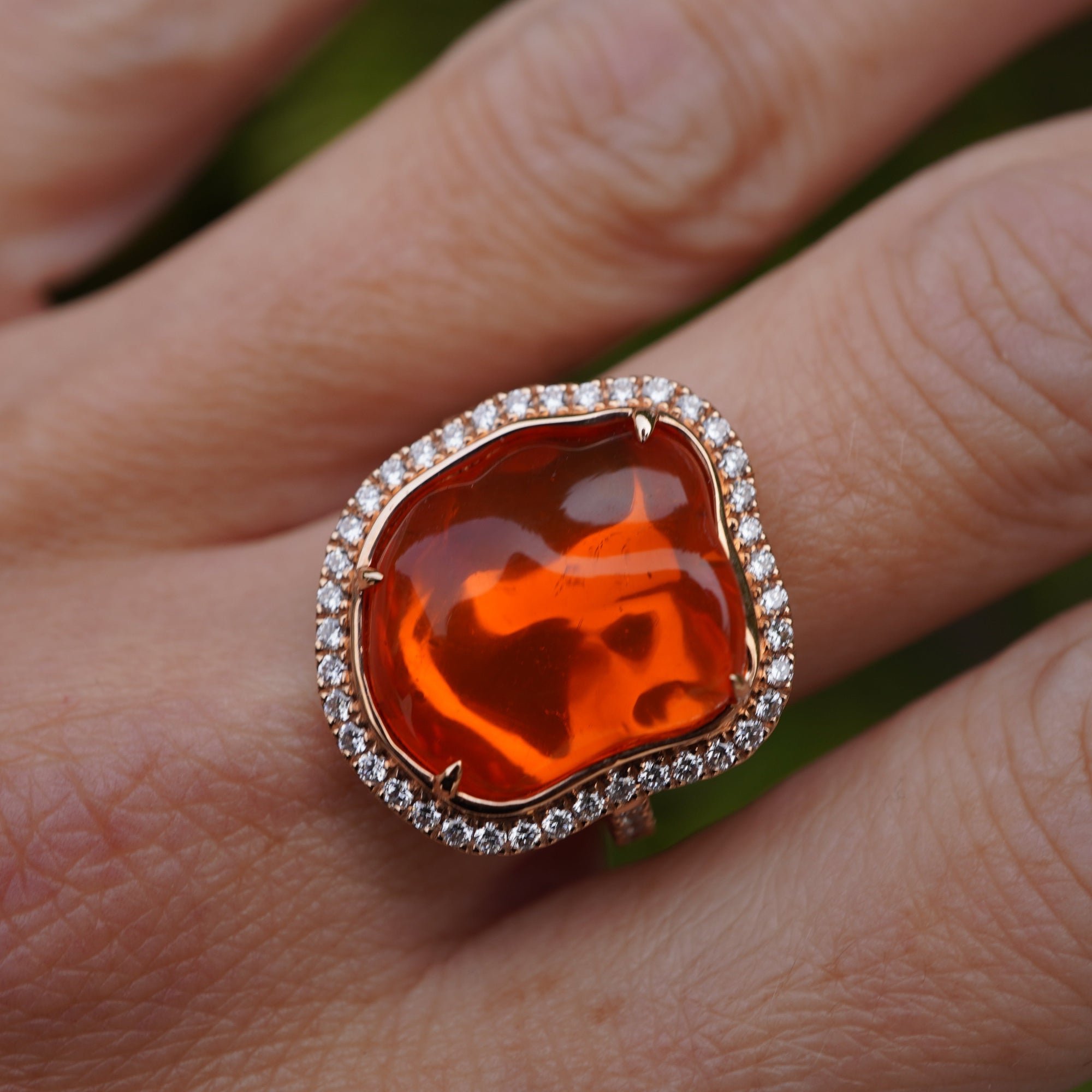 Captivating 18K rose gold ring featuring a glowing 6.21ct organic fire opal cabochon, encircled by a shimmering 0.51ct diamond halo for a radiant and enchanting statement piece. On models hand close up. 