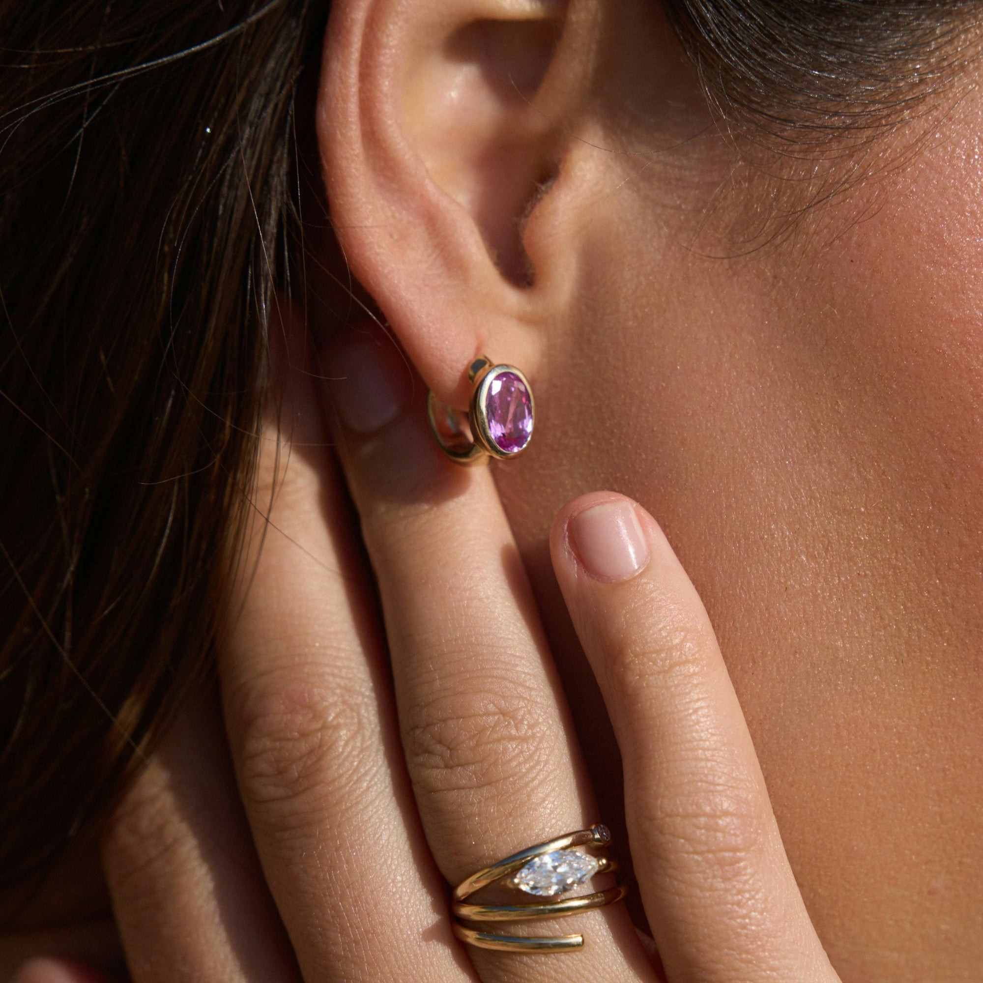 Enchanting pink sapphire huggie earrings crafted in 14K yellow gold, featuring vibrant oval-cut pink sapphires for a perfect blend of charm and sophistication, suitable for any occasion. On body, right ear. 